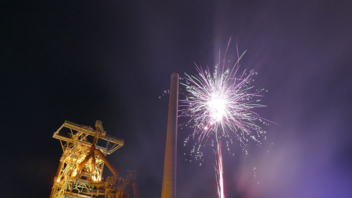 a photo at night. On the left is a lit up old steel structure from the coal mining times. In the centre a brick chimney, on the right side some fireworks 