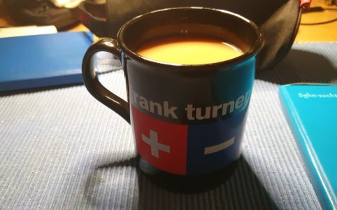 Coffee in a mug with a Frank Turner Logo on nit. A white plus sign on a red square, next to a white minus sign on a blue square.