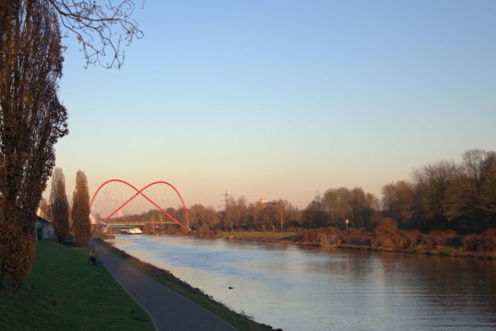 transportation canal with a towpath, in the lower third of the image. A unique red double arch foot bridge across the channel in the left middle. Blue sky with an evening tinge above.