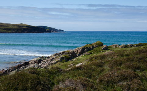 A turquoise ocean bay surrounded by green and rocky slopes in the foreground and the left side of the background. Clouds in the sky above on a sunny day