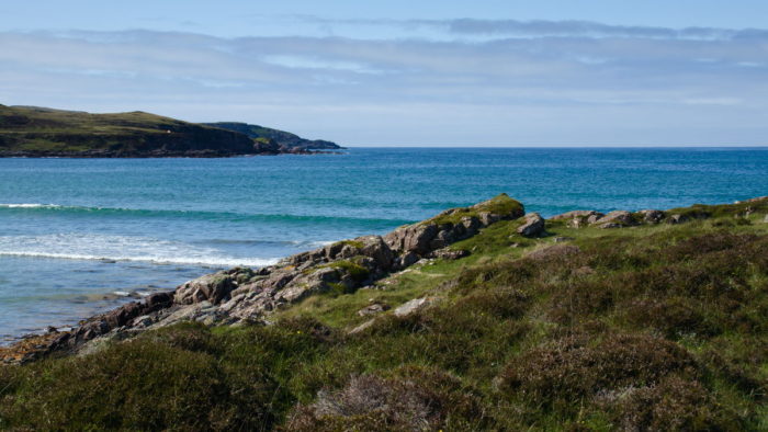 A turquoise ocean bay surrounded by green and rocky slopes in the foreground and the left side of the background. Clouds in the sky above on a sunny day