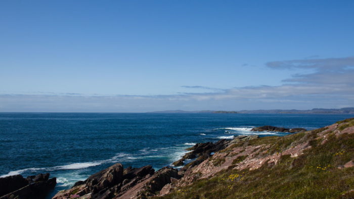 A turquoise ocean with green and rocky slopes in the foreground and some hills in the right background. Some clouds in the sky above on a sunny day