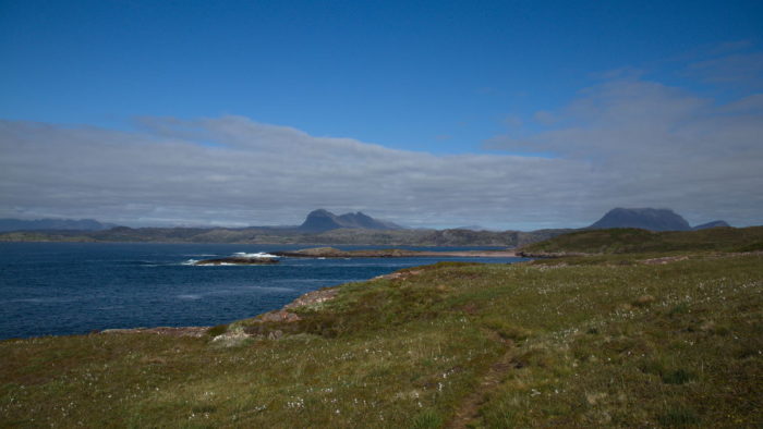 a view across an ocean bay with a green meadow in the foreground and two Scottish hills clearly visable across the bay in the background . Clouds in the sky above on a sunny day