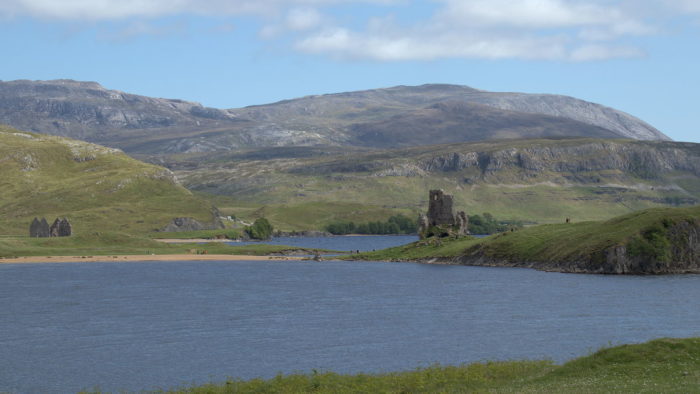 A loch in the forground, Adreck Castle ruins far away on the loch shore, hills and blue sky in the background