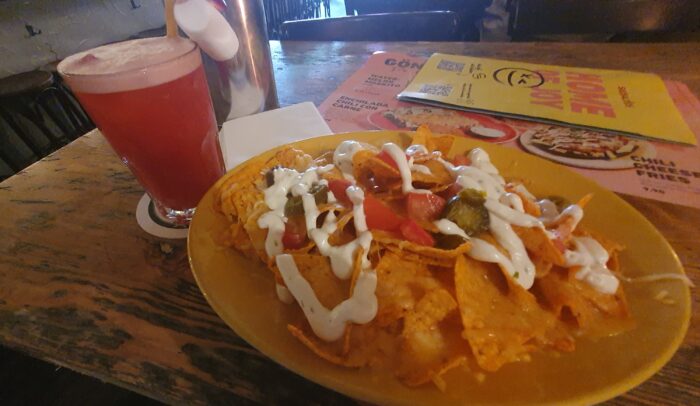 Red coloured cocktail and a plate of nachos with cheese, sour cream, tomato, jalapenos