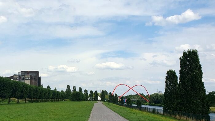 Old industrial building on the left, a canal with a unique red bridge on the right. Trees, meadow and a path straight ahead in the middle. Blue sky with clouds above