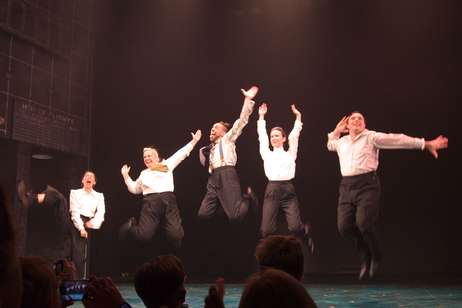 Picture shows three women, two men on stage before a black background. They are all wearing black trousers and white shirts. They are jumping up to various degrees