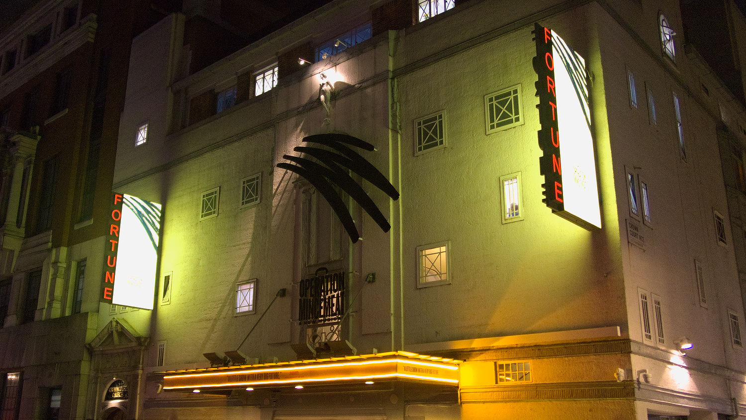 Picture shows the Fortune Theatre in London. The signs are lit in yellow. The front of the theatre has huge display of the Operation Mincemeat musical logo. Hitlers hair and below a smaller sign which looks like a suitcase: it says Operation Mincemeat a new musical