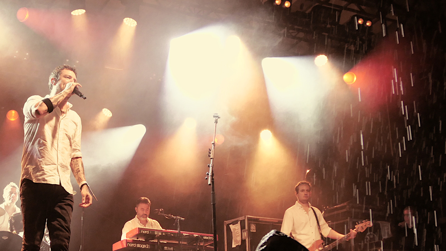 Photo of a stage. Frank Turner with just a mic, drummer Callum, on keys Matt and bass player Tarrant in the background. Heavy rain visible in the right side of the photo