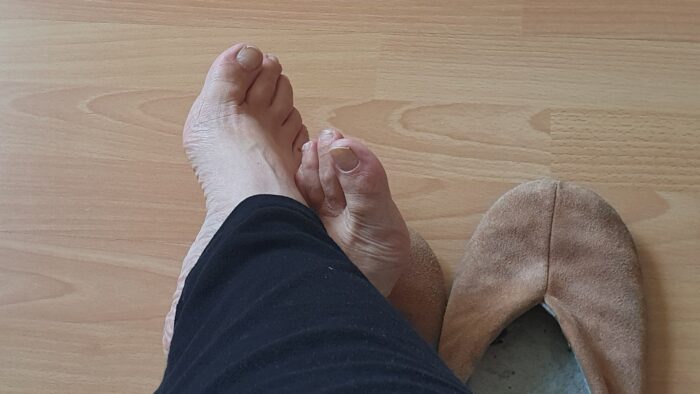 Photo of bare feet crossed over at the ankles on a wooden floor. Legs are cover in black trousers