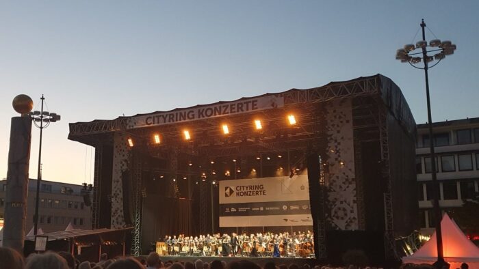 A big openair stage on a city plaza. On the stage is an orchestra, the sky is still a bit light, but dusky