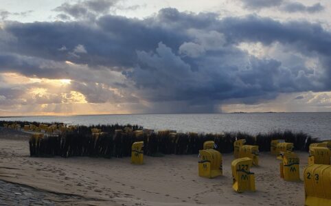 Photo of a beach at dusk. In the foreground yellow beach chairs on a beach, a still ocean in the middle. In the background the sun breaks through the clouds on the left while there are many dark clouds in the middle of the horizon
