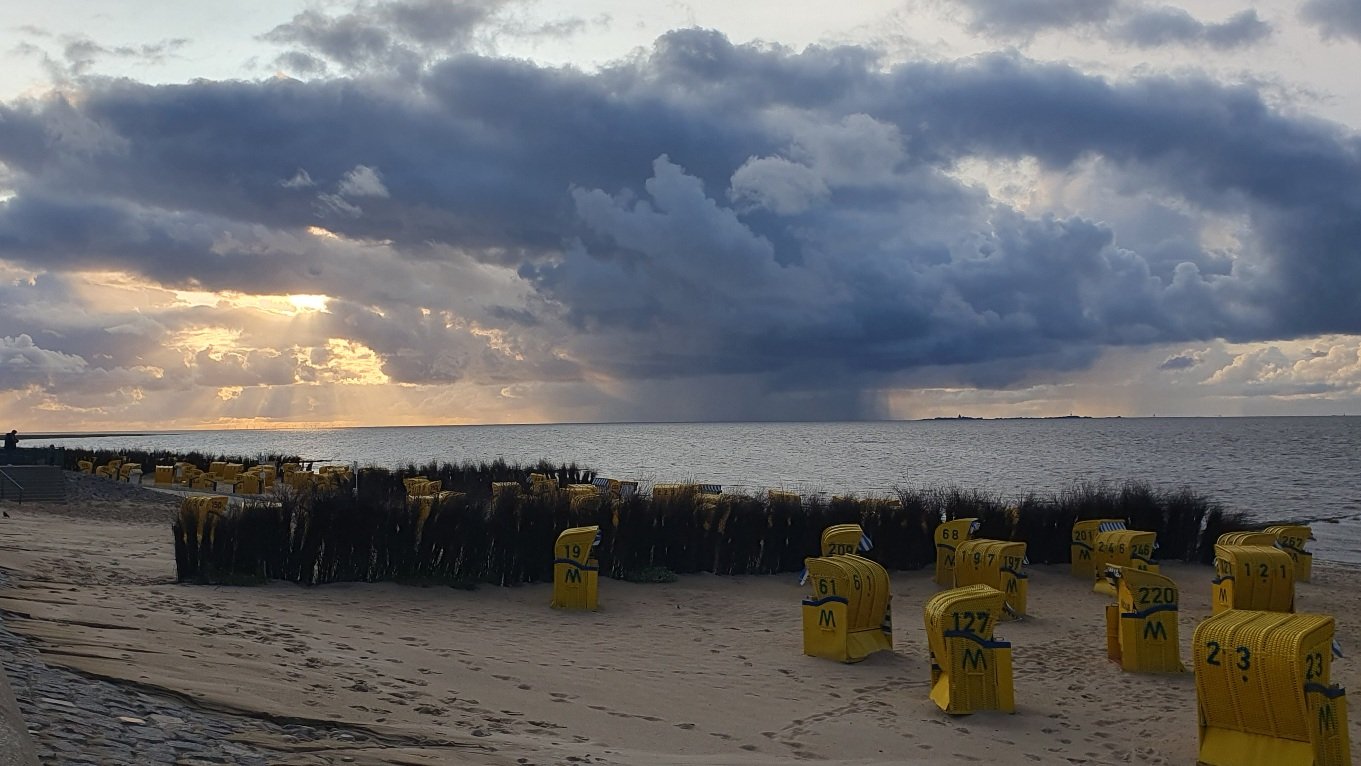 Photo of a beach at dusk. In the foreground yellow beach chairs on a beach, a still ocean in the middle. In the background the sun breaks through the clouds on the left while there are many dark clouds in the middle of the horizon