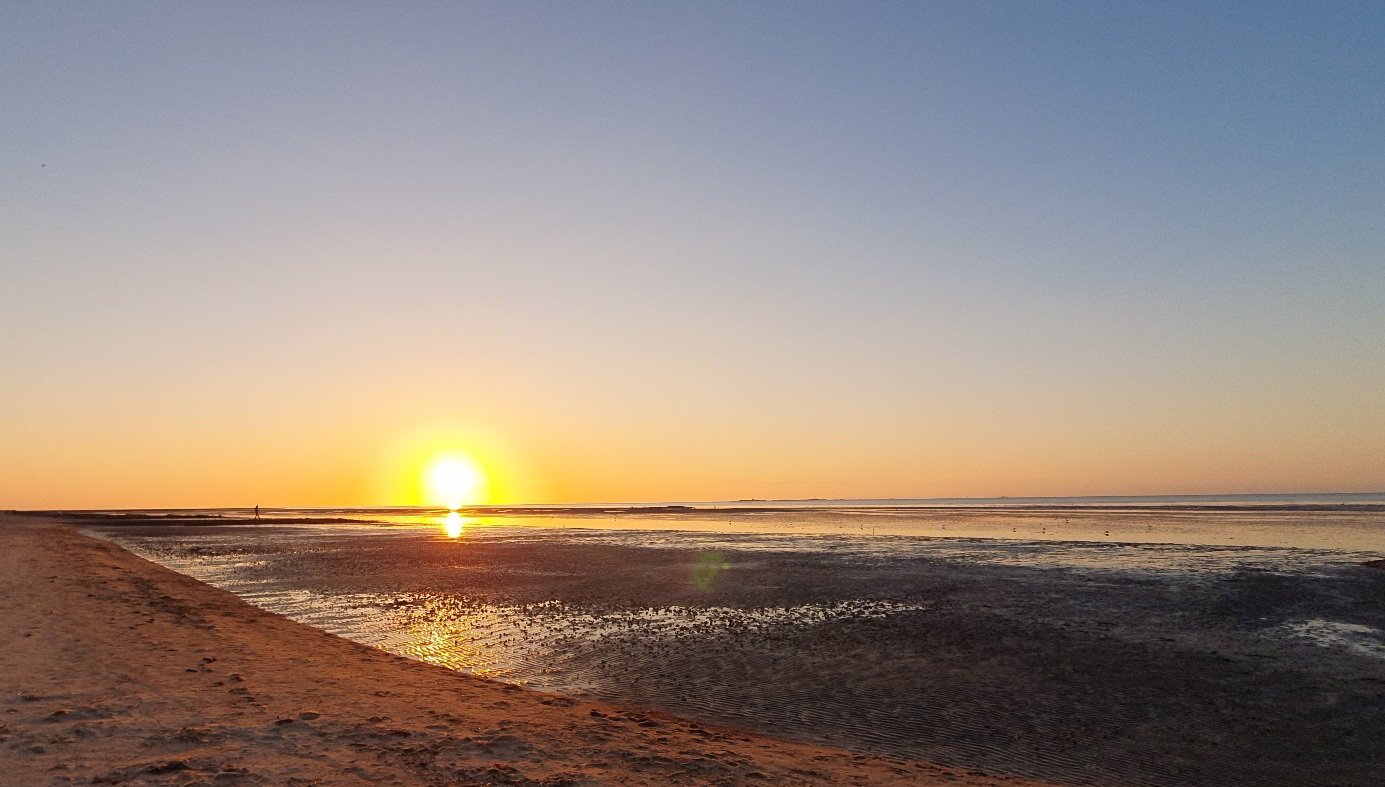 Photo of a sunset at a beach with lowtide. Sandy beach on the left side, low tide in the middle and right. The sun sets in the corner of the lower left quadrant. Cloudless sky
