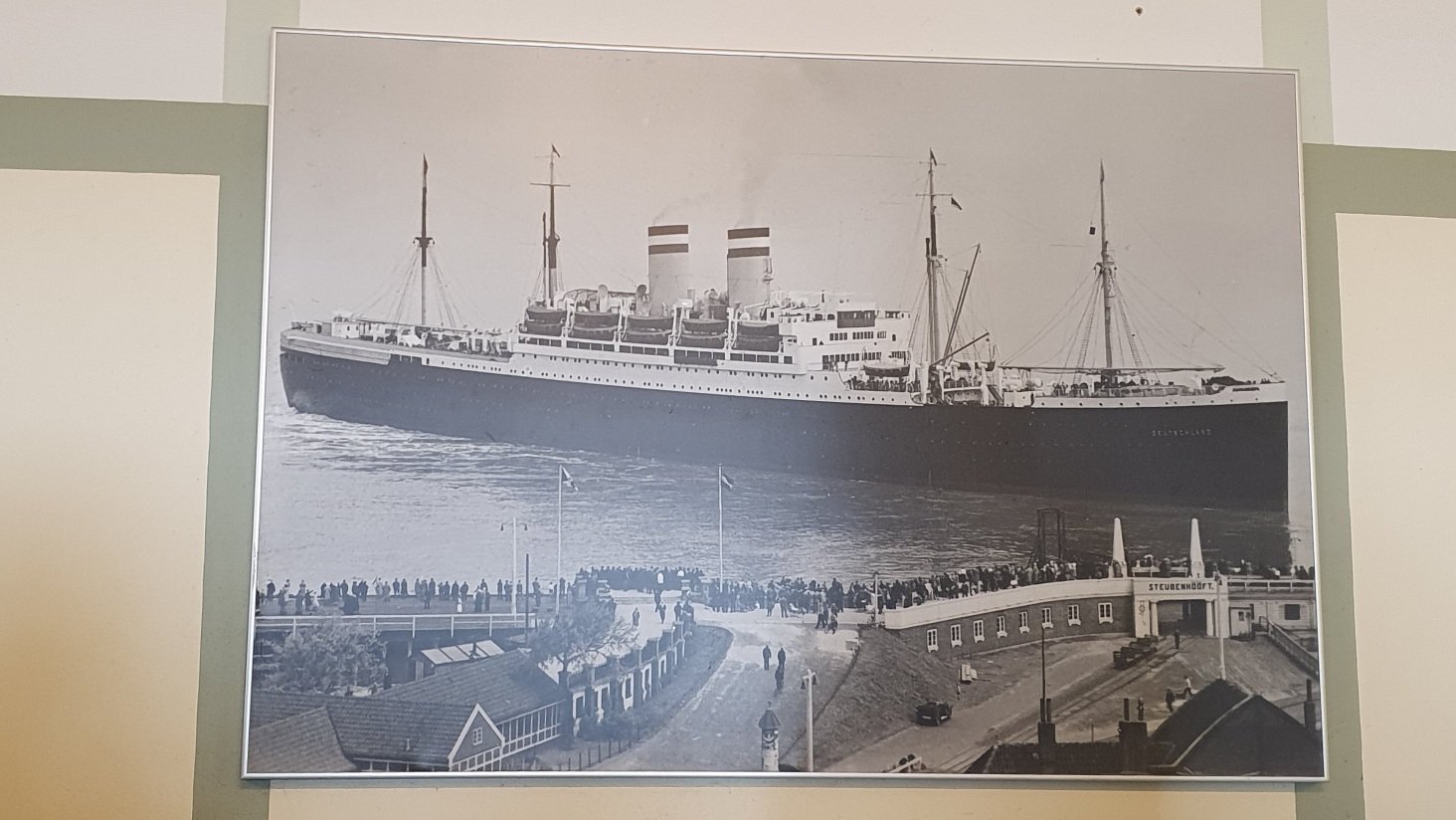 Photo of large black and white photo from mid 20th century. A big ocean liner, in the foreground a pier of people