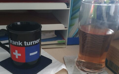 Photo of a desk with file drawers in the background. Centre are a black mug with a Frank Turner print and a pint glass, half full of a peach coloured liquid