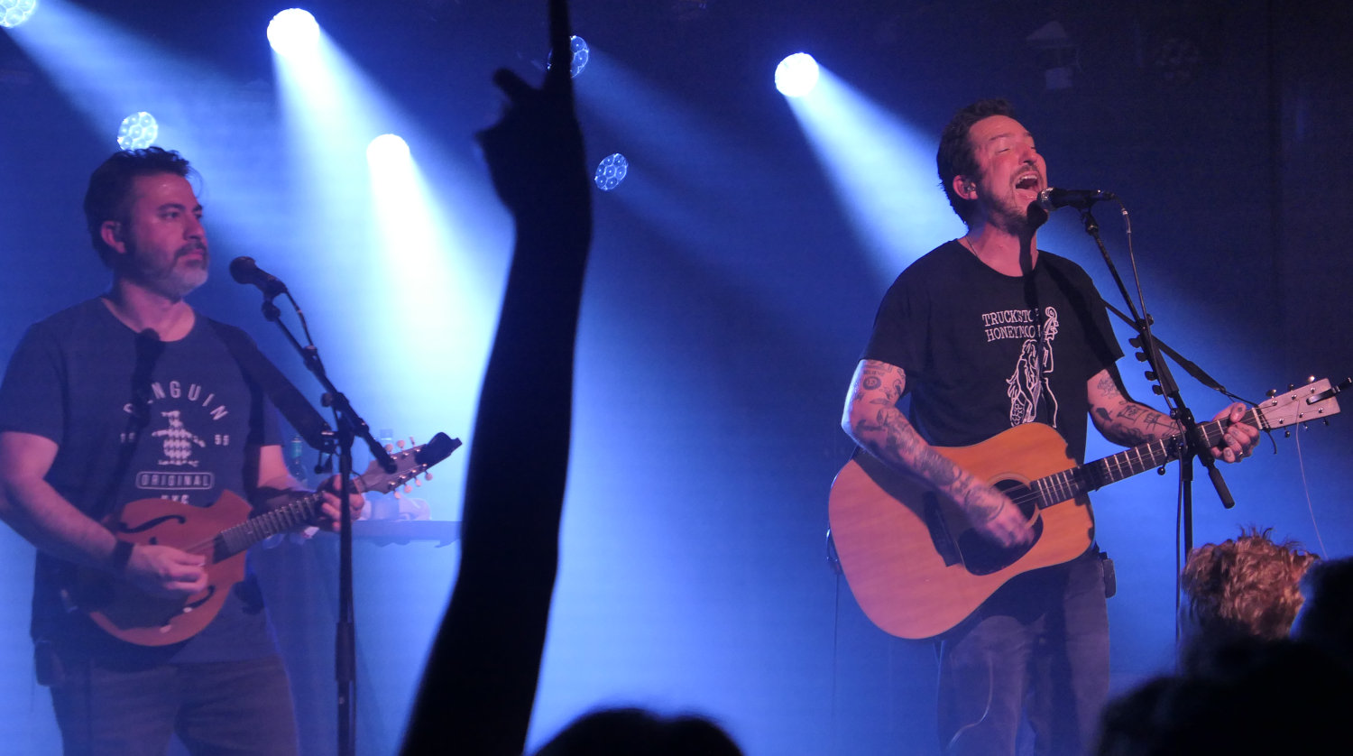 Photo of Matt Nasir on mandoline and Frank Turner with a guitar on stage. In the middle foreground is a raised arm in the shadow