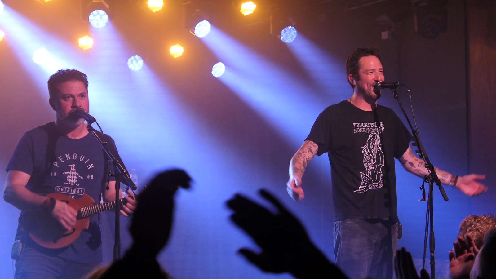 Photo of Matt Nasir on mandoline and Frank Turner without a guitar on stage. Frank is smiling. In the foreground there are a few clapping hands