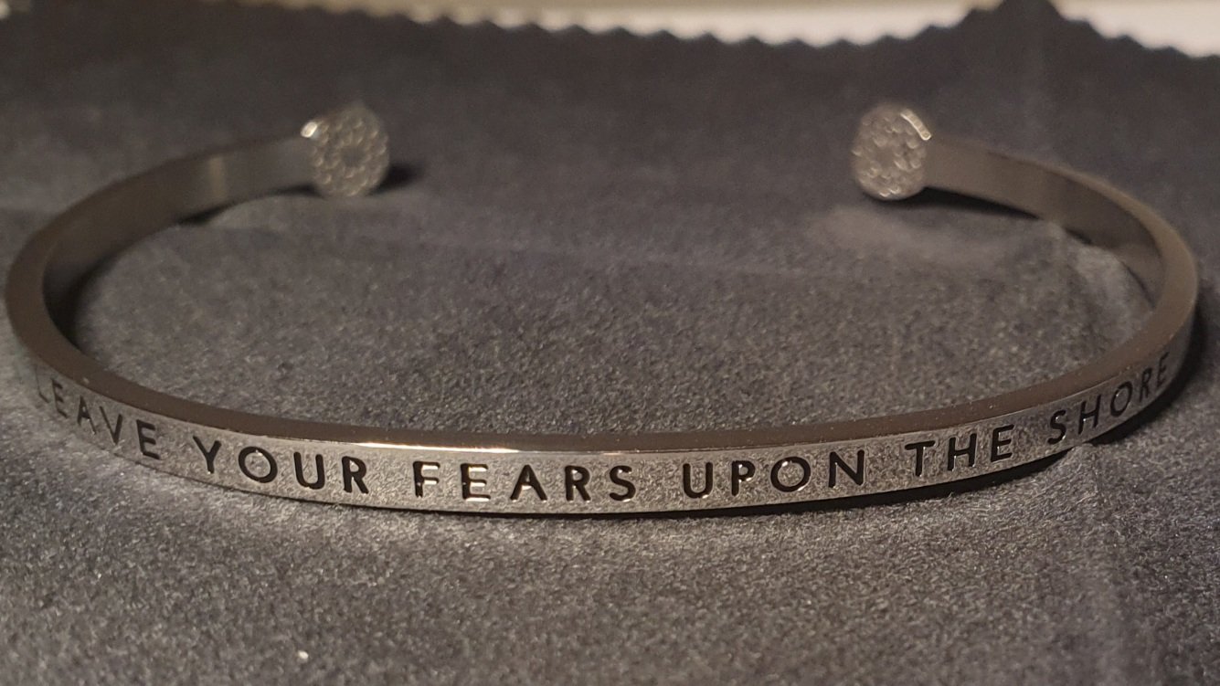 Photo of a silver bracelet on a black cloth. Bracelet inscribed with "leave your fears upon the shore" in capital letters
