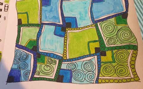 Photo of a half-finished doodling. Wonky squares filled in shades of blue and green. circles drawn on the border lines and spirals in some of the squares