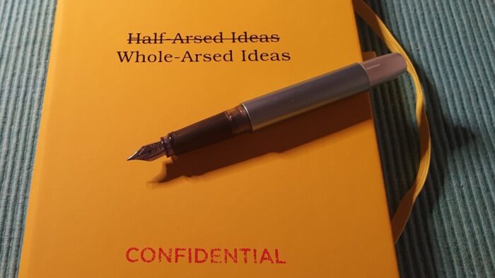 Photo of a yellow notebook with the words whole-arsed ideas printed on it. A open fountain pen lies on the cover