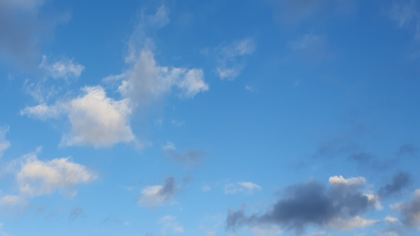 Photo of a blue sky with a few white clouds and one grey one in the bottom right corner