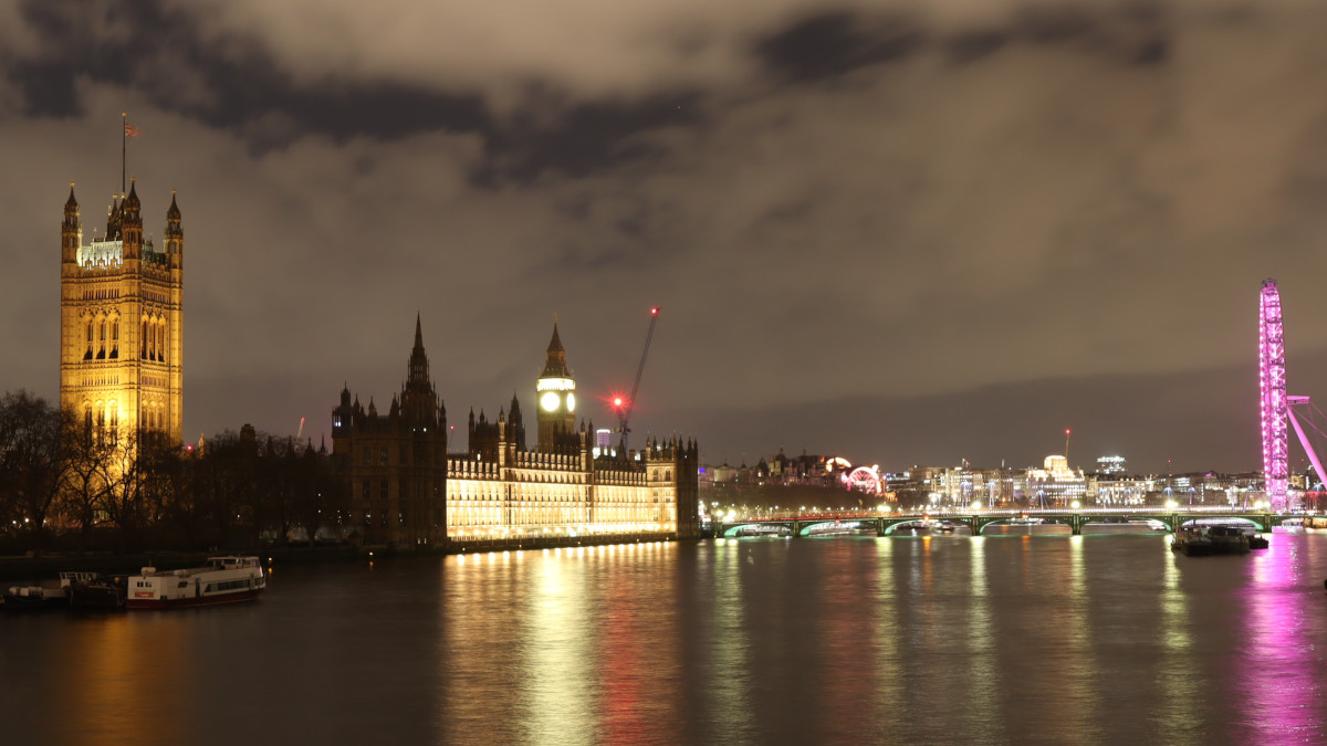 Photo of Westminster London by night. On the right the House of Parliament on the right the London Eye in pink light. Westminster bridge and more of central London in the centre. Clouds in the sky