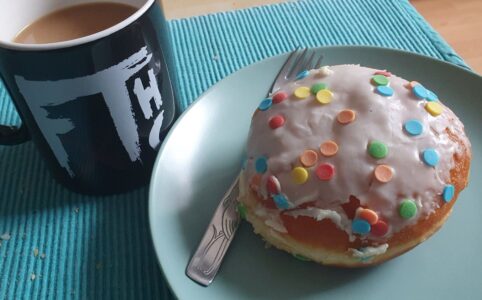 Black mug with FTHC (Album) logo, filled with coffee. Next is a Berliner (pastry) with sugar confetti icing on a green plate