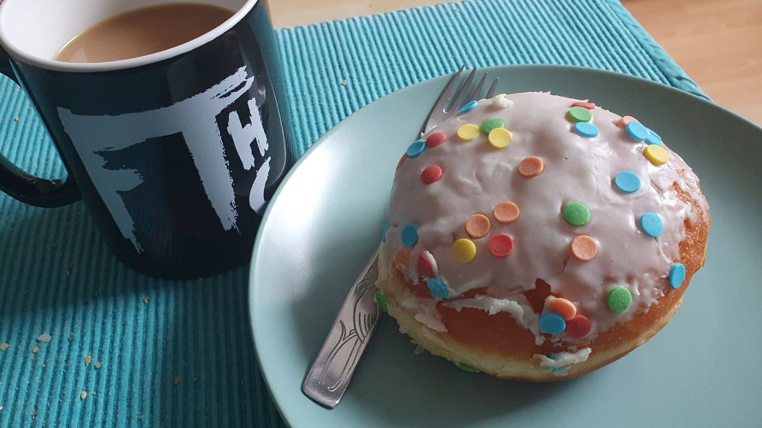 Black mug with FTHC (Album) logo, filled with coffee. Next is a Berliner (pastry) with sugar confetti icing on a green plate