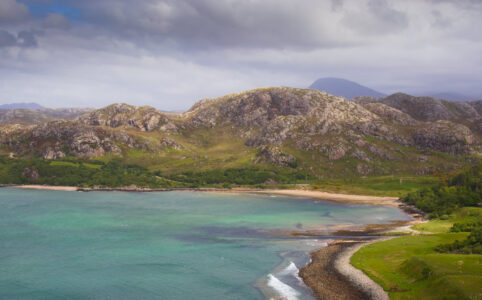 A bay with turquoise water in the lower half of the image, on the right side a bit of beach an green meadows. Scottish granite hills in the centre of the image and cloudy sky above