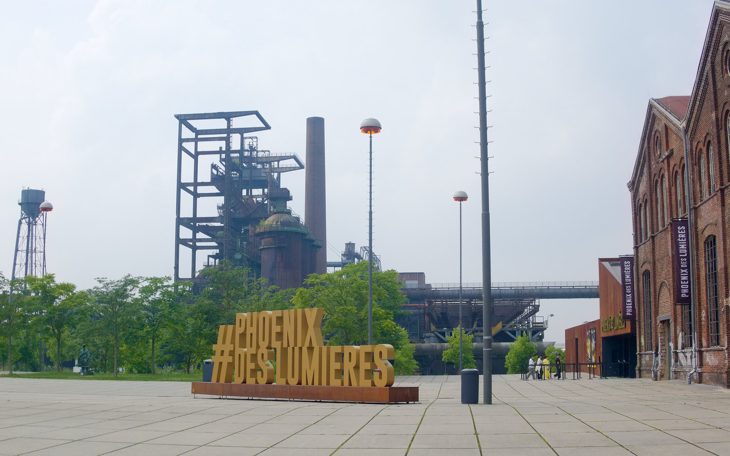 Phoenix des Lumieres sign on the square in front of a brick building with former industrial iron buildings in the background