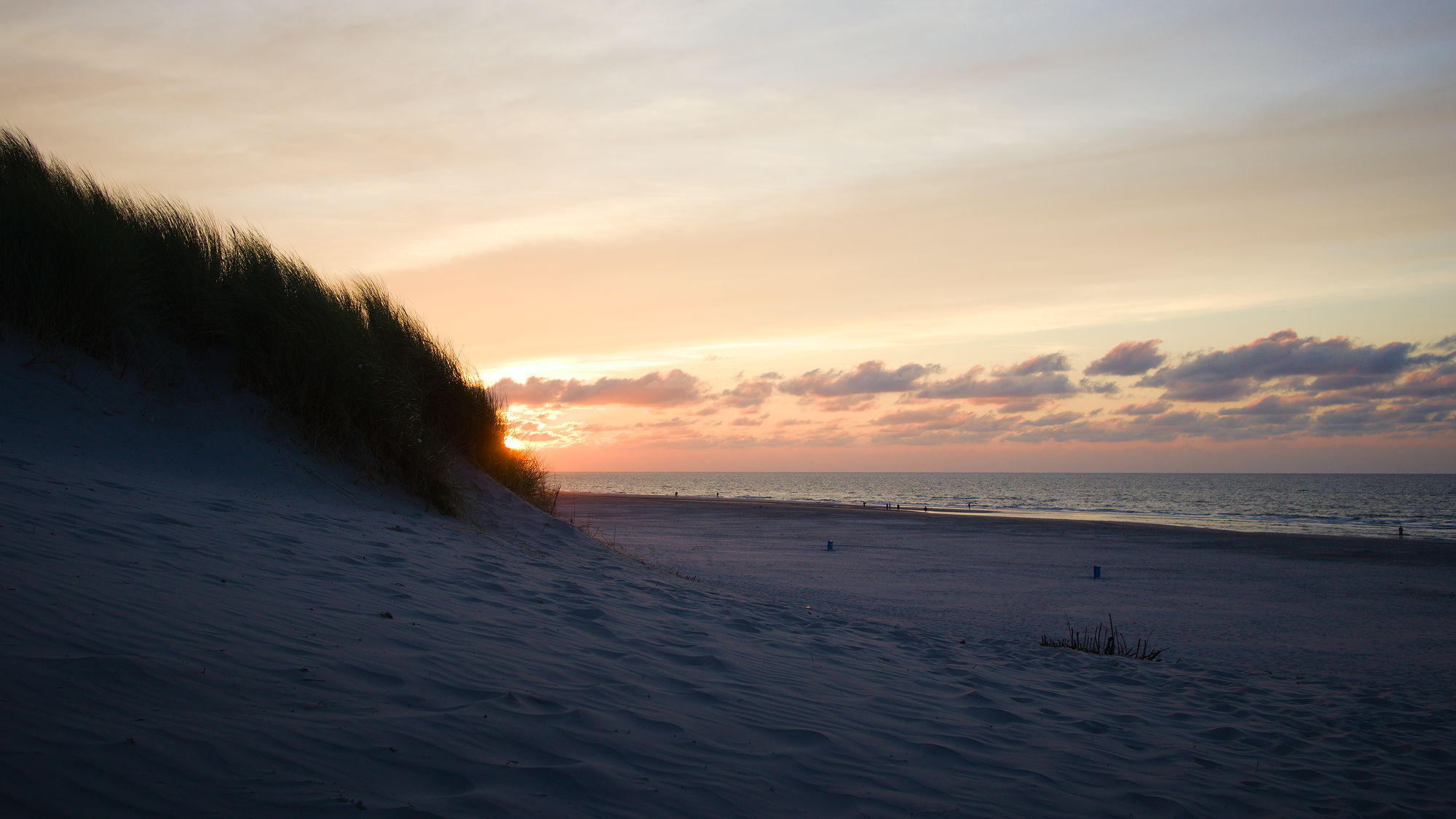 A dune with grass on the right side, the wide beach in the middle of the image, bordering on the ocean. The sun sets behind the dunes, the sky is a bit cloudy and dusky