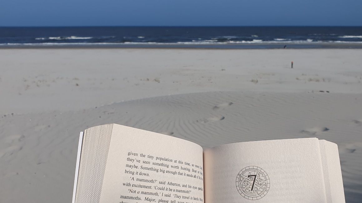 Close up of half a page of a paperback in focus, beach and the ocean blurry in the background