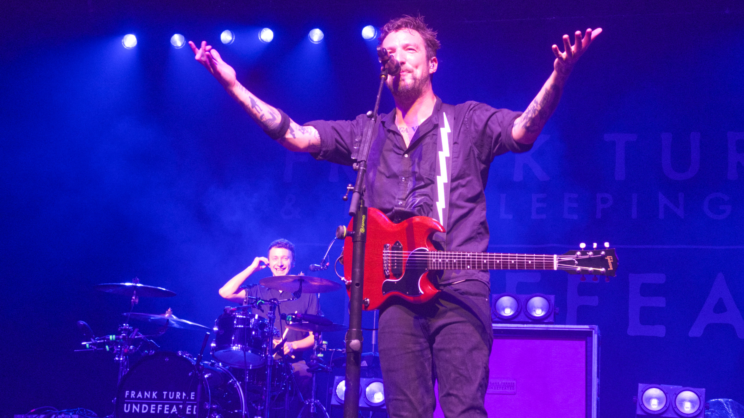 Frank Turner on stage, Callum Green on drums in the background smiling