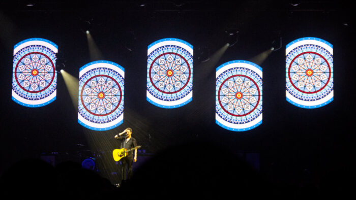 Frank Turner solo on stage with 5 images of the Alexandra Palace stained glass window behind him