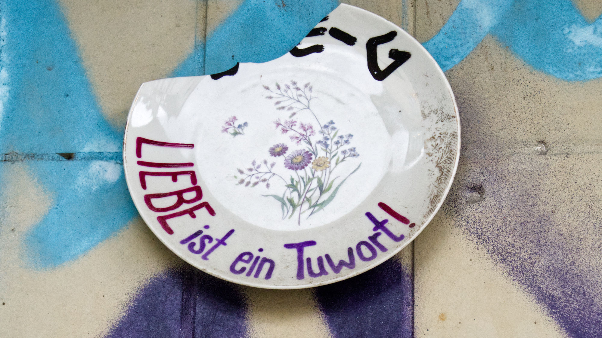 Broken plate on a wall, written on it "Liebe ist ein Tuwort" (Love is a verb)